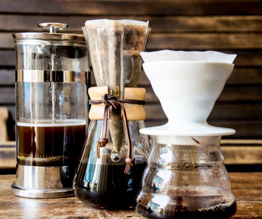 En la imagen hay tres cafeteras: una prensa francesa de vidrio con émbolo metálico, una cafetera eléctrica por goteo con jarra de vidrio, y una cafetera pour-over manual con un cono sobre una taza. Están alineadas sobre una encimera.
