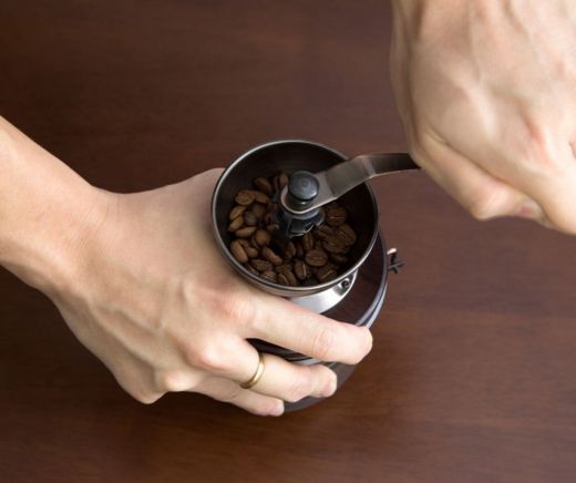 Molino manual que contiene granos de cafe mientras un par de manos lo trabajan